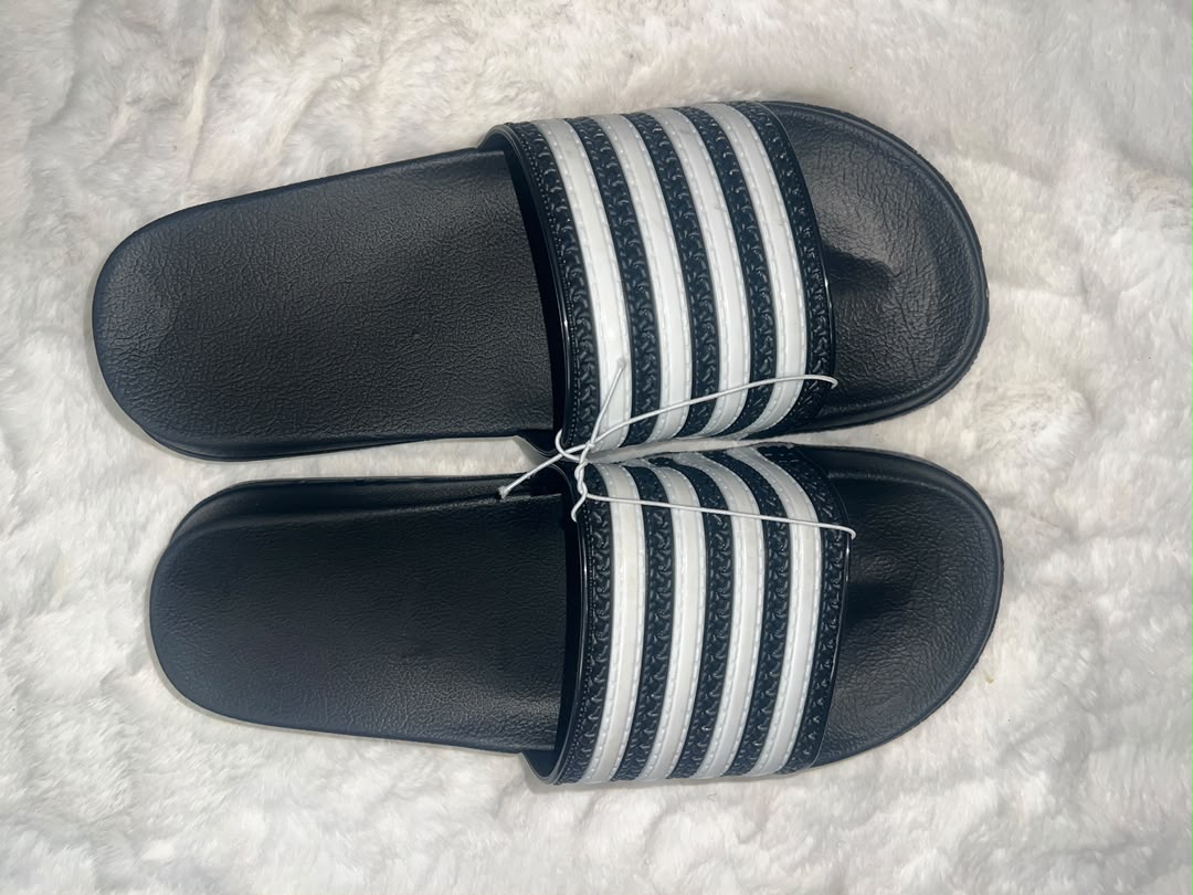 A pair of Adidas slide sandals with black footbeds and white stripes on the straps, laid flat on a white textured rug.