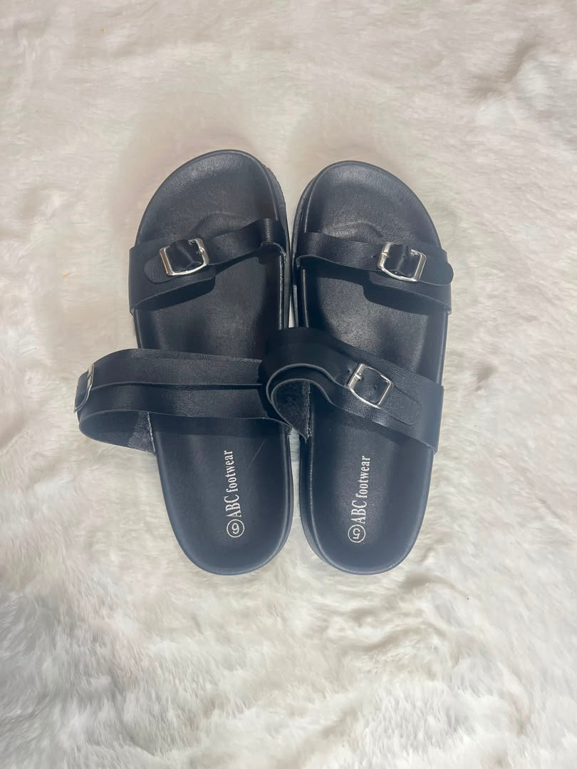 A pair of black slide sandals with double straps and silver buckle details, displayed on a white faux fur surface. The brand name 'ABC Couchwear' is printed on the soles.