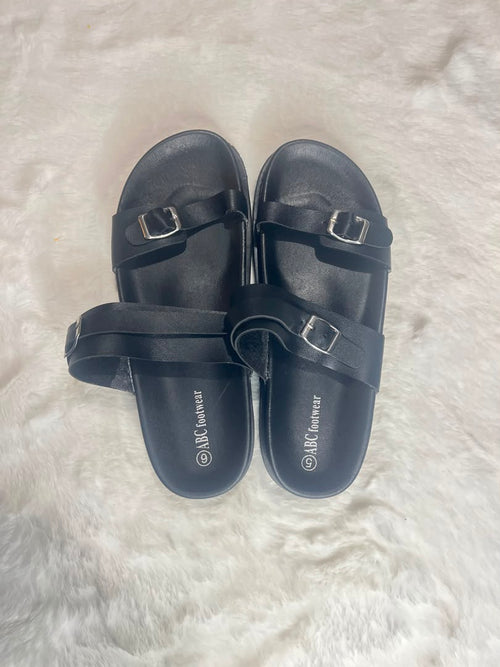 A pair of black slide sandals with double straps and silver buckle details, displayed on a white faux fur surface. The brand name 'ABC Couchwear' is printed on the soles.