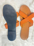 A pair of tan leather slide sandals shown from above on a white faux fur background, with one sandal displaying the textured dark gray outsole and the other showing the tan footbed and orange-brown straps.
