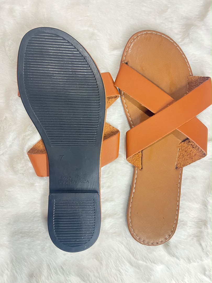 A pair of tan leather slide sandals shown from above on a white faux fur background, with one sandal displaying the textured dark gray outsole and the other showing the tan footbed and orange-brown straps.