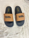 A pair of Zara slide sandals with black footbeds and textured tan straps featuring a raised black ZARA logo, displayed on a white faux fur surface.