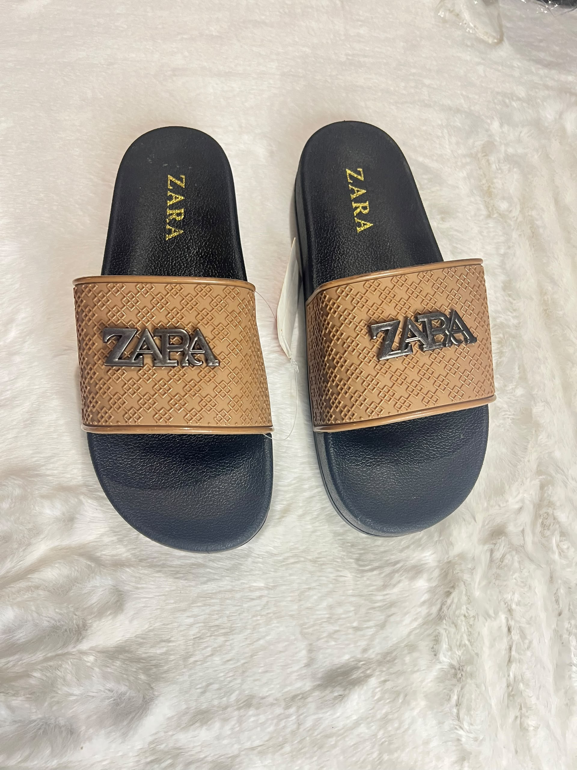 A pair of Zara slide sandals with black footbeds and textured tan straps featuring a raised black ZARA logo, displayed on a white faux fur surface.