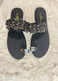 A pair of black flat thong sandals with a wide embellished strap covered in black and silver rhinestones, and matching silver toe rings, displayed on a white faux fur background.