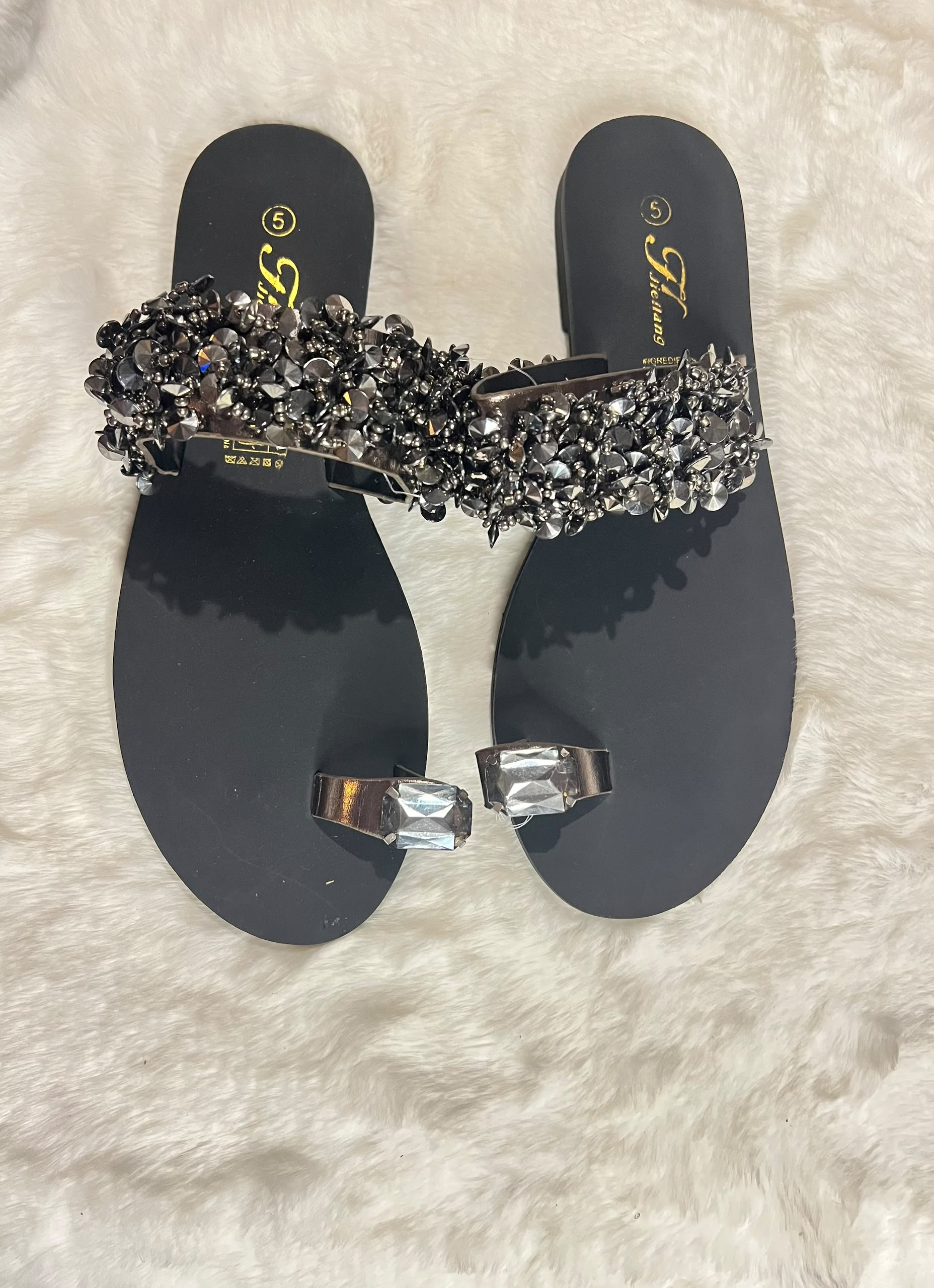 A pair of black flat thong sandals with a wide embellished strap covered in black and silver rhinestones, and matching silver toe rings, displayed on a white faux fur background.