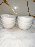 Four small white ceramic bowls, two stacked on the left and two stacked on the right, placed on a white textured surface with a white marble-patterned background.