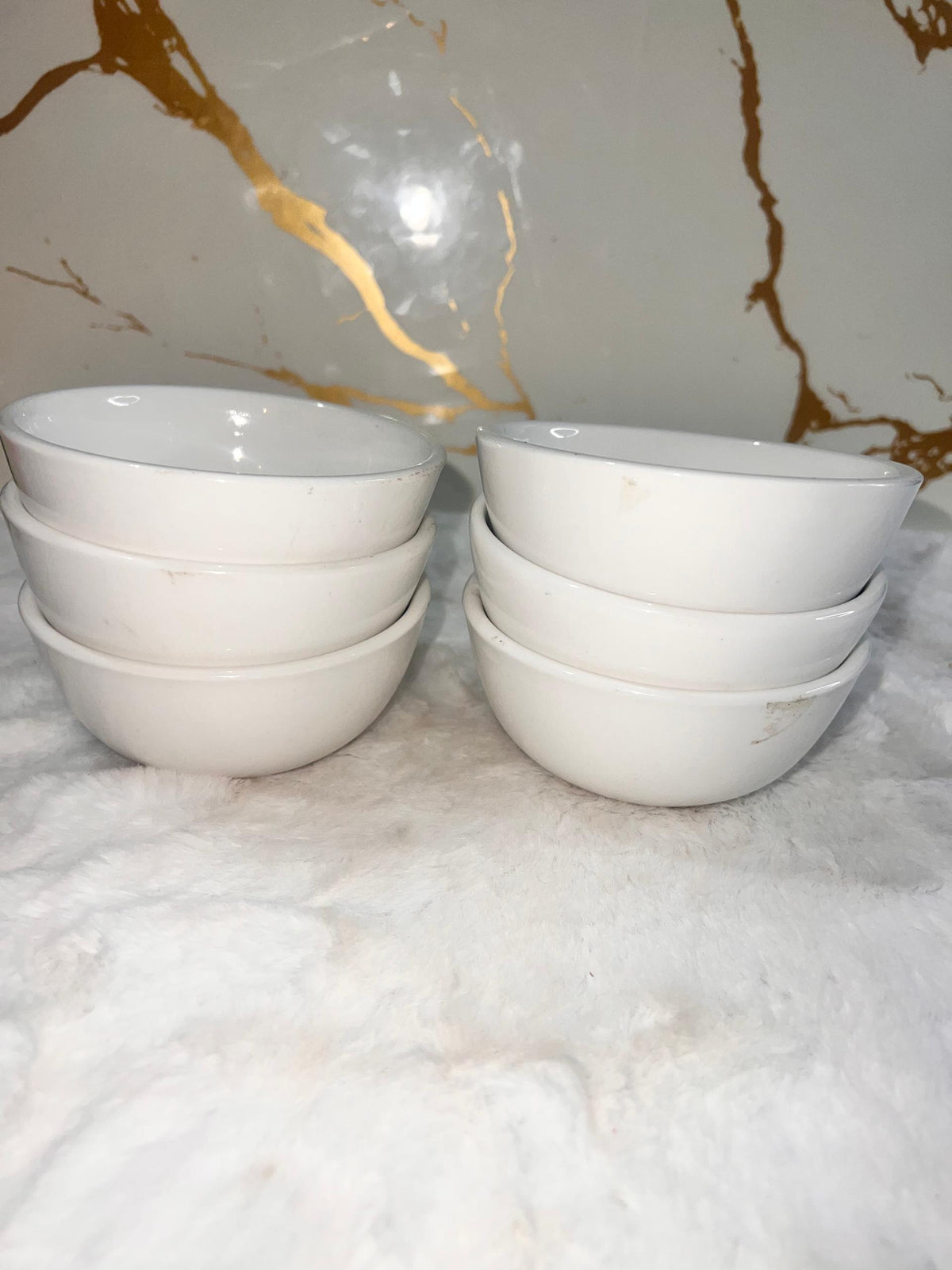 Four small white ceramic bowls, two stacked on the left and two stacked on the right, placed on a white textured surface with a white marble-patterned background.