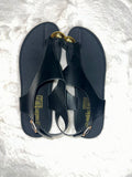 A pair of black flat T-strap sandals with gold-tone decorative accents, displayed on a white textured faux fur surface. The brand name 'sadlike' is printed in gold on the insole.