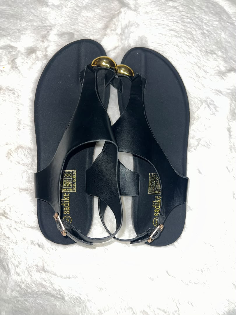 A pair of black flat T-strap sandals with gold-tone decorative accents, displayed on a white textured faux fur surface. The brand name 'sadlike' is printed in gold on the insole.