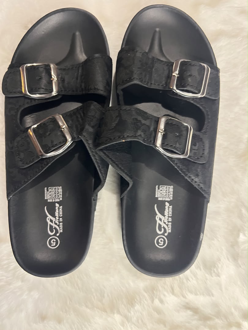 A pair of black double-strap slide sandals with silver buckle closures, laid flat on a white faux fur surface. The brand logo 'G' with 'Genting' is visible on the footbed.