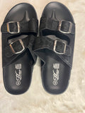 A pair of black double-strap slide sandals with silver buckle closures, laid flat on a white faux fur surface. The brand logo 'G' with 'Genting' is visible on the footbed.