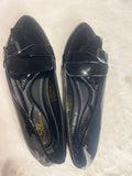 A pair of black patent leather loafers with a decorative bow detail across the vamp, displayed on a white faux fur surface. The brand name 'SARAH' is visible in gold lettering on the insole.