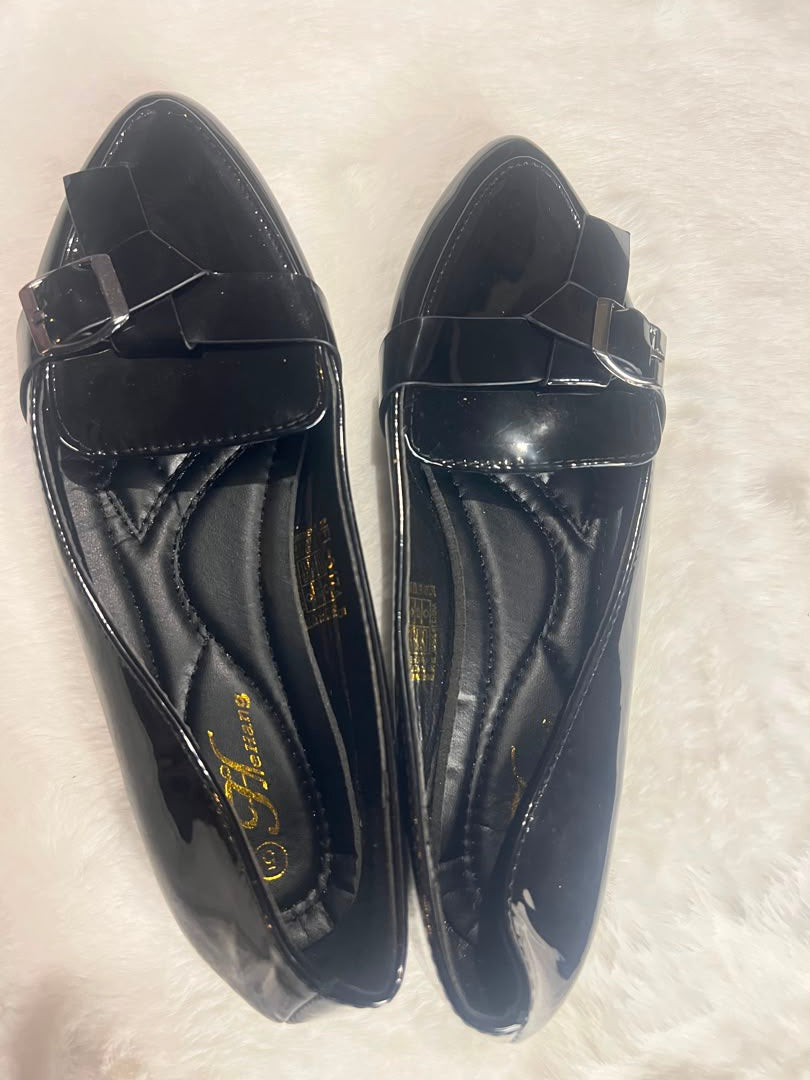 A pair of black patent leather loafers with a decorative bow detail across the vamp, displayed on a white faux fur surface. The brand name 'SARAH' is visible in gold lettering on the insole.