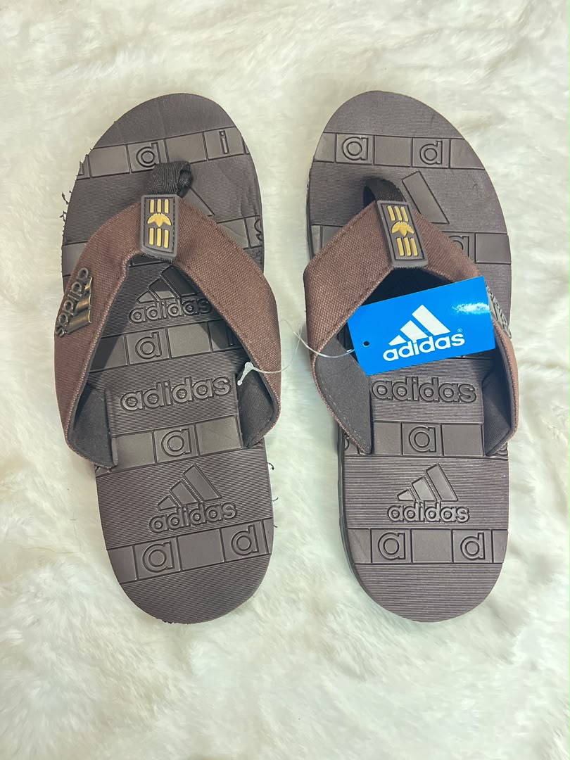 A pair of Adidas slide sandals in brown and dark gray, displayed flat on a white faux fur surface. One sandal has its blue retail tag attached.