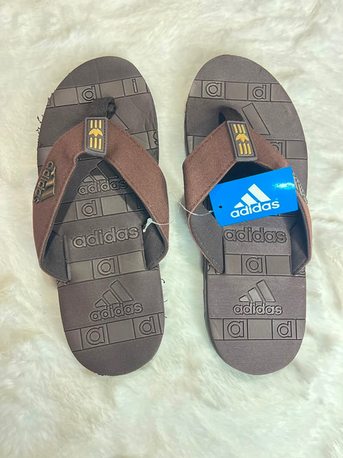 A pair of Adidas slide sandals in brown and dark gray, displayed flat on a white faux fur surface. One sandal has its blue retail tag attached.