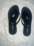 A pair of black flip-flop sandals with rhinestone-embellished straps, displayed on a white textured faux fur surface. The brand name 'Sadike' is printed on the bottom of the soles.